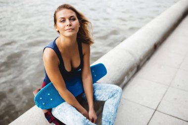 kız kameraya bakarak yol sınırındaki nehir kıyısında oturan mavi kaykay ile