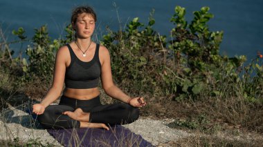 Yoga minderinde oturan ve deniz kıyısında lotus pozisyonunda meditasyon yapan genç bir kadın.