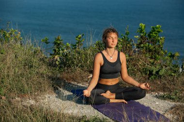 Yoga minderinde oturmuş deniz kıyısında meditasyon yapan gözleri kapalı genç bir kadın.
