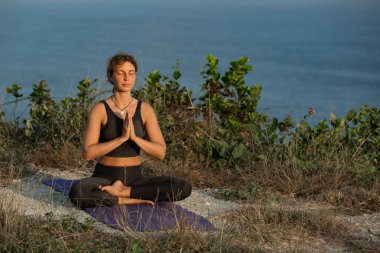 Yoga minderinde oturmuş okyanus kenarında meditasyon yapan genç bir kadın.
