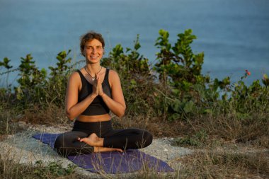 Genç bir kadın dışarıda yoga yaparken lotus pozisyonunda oturuyor ve kameraya gülümsüyor. 