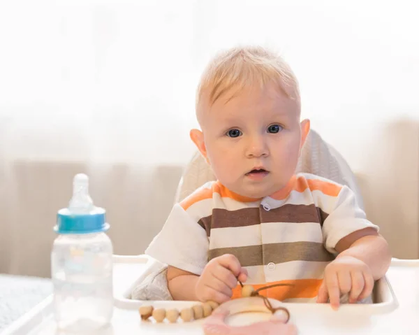 A small surprised blue-eyed child sits at a table and looks at the camera. On the table is a bottle with a pacifier and water. Amazement