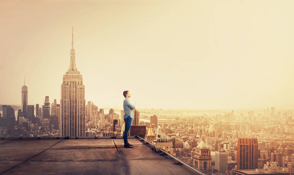 Side view of a thoughtful young businessman standing on concrete rooftop of a skyscraper watching over the big city at sunset. Business research concept.