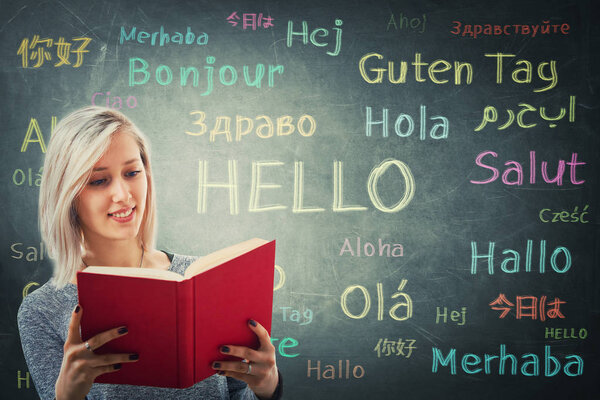Pretty girl holding a red book in front of a huge blackboard written with the word hello in different languages and colors. Opportunity for learning many languages for students.