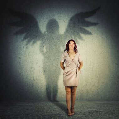 Funny young woman hiding hands behind her back looking away. Full length female portrait casting a superhero shadow with angel wings on a dark room wall. Inner power, ambition and leadership concept. clipart