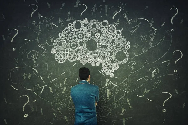 Rear view of a puzzled businessman having questions in front of a blackboard. Big chalk drawn gear brain above head, positive thinking mess as thoughts. Concept for mental, psychological development.
