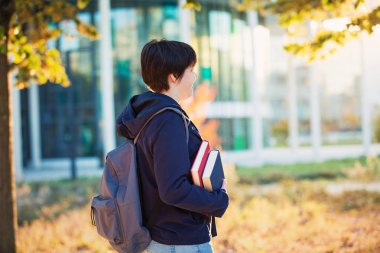 Yan görünümü açık havada genç öğrenci kız portresi iki kitap tutan ve taşıyan üniversiteye güneşli sonbahar günü yürüyüş gitmek sırt çantası.
