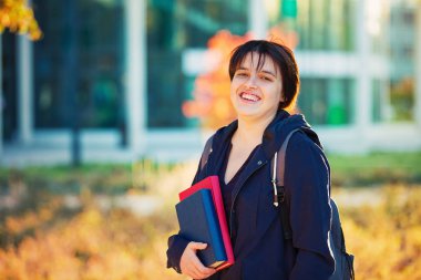 Neşeli kadın öğrenci kitap ve çantası elinde kamera seyir gülümseyerek. Açık havada sonbahar portre, üniversite kampüsü.