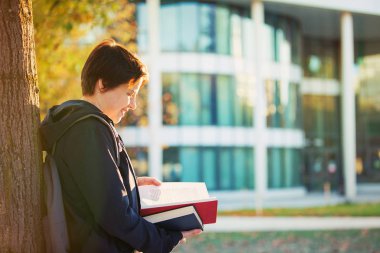 Yan görünümü açık havada sonbahar portresi kampüs geri bir ağaç gövdesi ve bir kitap okuma yağsız bir genç kadın öğrenci.