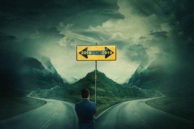 Man in front of a crossroad, fork junction and road sign arrows splitted in two different ways. Go back in 2018 or move to 2019 year. Choosing correct pathway. Live in the past or change your life. clipart