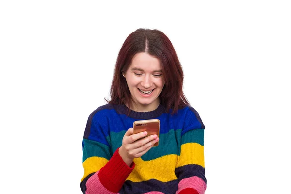 愉快的休闲年轻女孩五颜六色的毛衣举行智能手机浏览互联网与她的脸上的笑容 积极的情绪孤立在白色的背景 — 图库照片