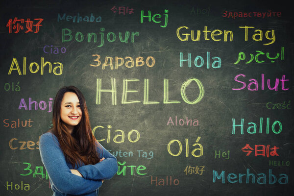 Cheerful woman student holding arms crossed in front of a huge blackboard written with the word hello translated in different languages and colors. Opportunity for learning and speaking a language.
