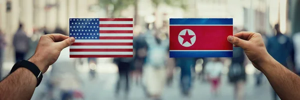 Two hands holding different flags, USA vs North Korea Republic on politics arena over crowded street background. Future strategy, relations between countries. Cooperation or opposite conflict concept.