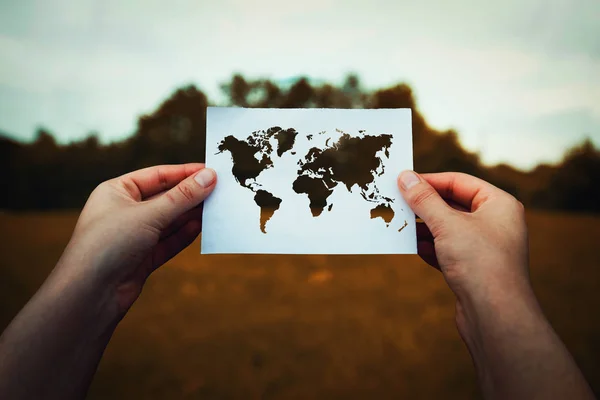 Climate change problem, destruction of nature concept. Human hands holding a paper sheet with world map icon over a dry grass field background. Global warming, solar radiation and pollution risk.