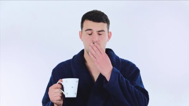 眠いカジュアルな若者がバスローブを着て 朝にコーヒーや紅茶を飲みながら白い背景に孤立したテレビチャンネルを切り替える — ストック動画