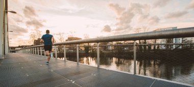 Genç atletik adam dışarıda spor yapıyor, günbatımı gökyüzü arka planında köprü boyunca hızla koşuyor. Benlik fetheden engelleri aşar ve kazanır. Sağlıklı yaşam tarzı kavramı. Koşu etkinliği. Panoramik pankart