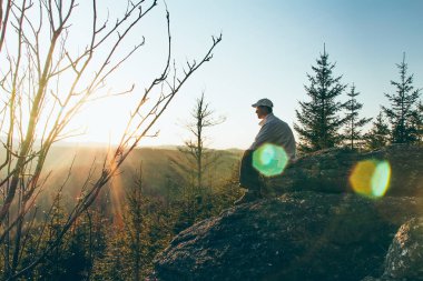 Young man hiker in white cap and shirt sit on rock from side wit