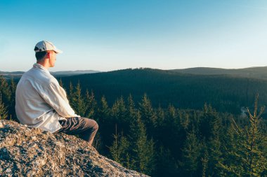 Young man hiker in white cap and shirt sit on rock from back loo
