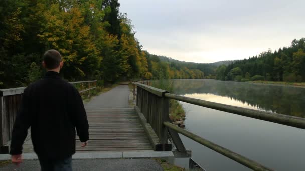 Молодой человек ходит по деревянному мосту с рекой Влтавой и осенью — стоковое видео