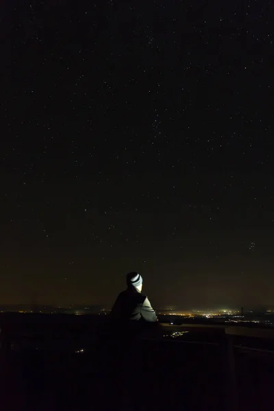 Молодой человек смотрит на ночное небо, полное звезд с далеким городом , — стоковое фото
