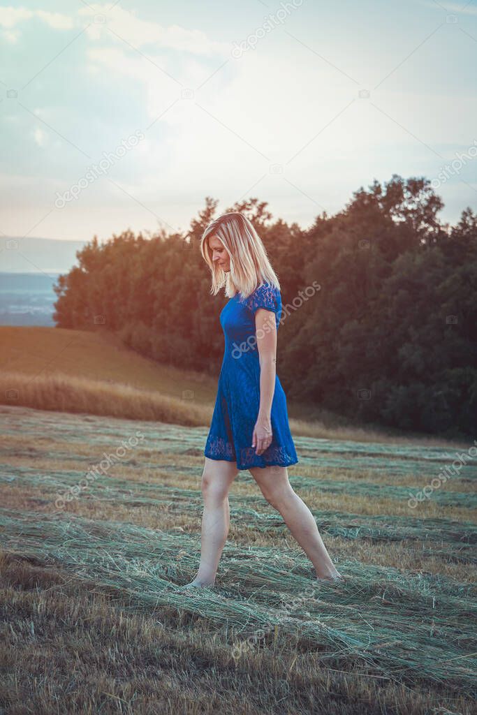 Beauty blonde hair young woman in blue dress walking in mowed grass. Muted colors