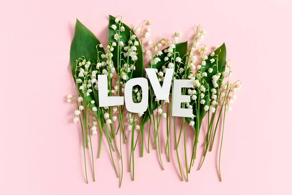 Word Love made of letters cut out of paper. Lilies of the valley and green leaves on a pink pastel background
