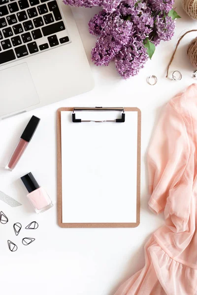 Workspace with clipboard, laptop, pink dress, bouquet of lilac, cosmetics and accessories