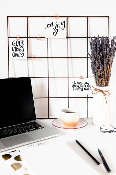 White workspace with quotes on mood board, laptop, stationery, mug of coffee and bouquet of lavender flowers