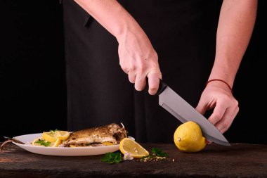 kadın mutfakta yemek için ahşap tahta üzerinde pişmiş balık elinde görüntülemek için limon kesme. Limon, baharatlar ve otlar beyaz plaka üzerinde pişmiş balık