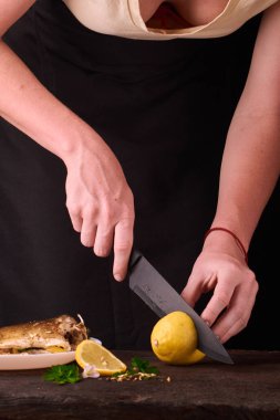 kadın mutfakta yemek için ahşap tahta üzerinde pişmiş balık elinde görüntülemek için limon kesme. Limon, baharatlar ve otlar beyaz plaka üzerinde pişmiş balık