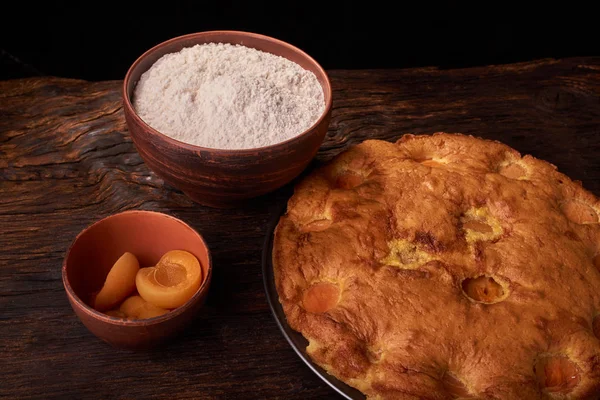 A bowl with flour and apricot cake on a wooden background on black