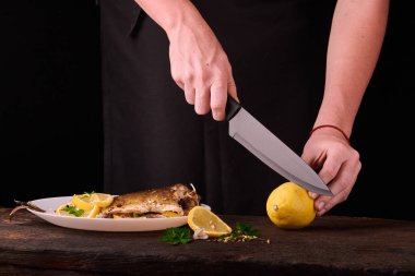kadın mutfakta yemek için ahşap tahta üzerinde pişmiş balık elinde görüntülemek için limon kesme. Limon, baharatlar ve otlar beyaz plaka üzerinde pişmiş balık