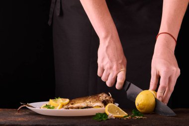 kadın mutfakta yemek için ahşap tahta üzerinde pişmiş balık elinde görüntülemek için limon kesme. Limon, baharatlar ve otlar beyaz plaka üzerinde pişmiş balık