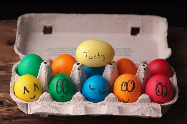Colorful Easter eggs with painted face in egg carton box on wooden background