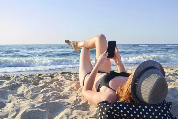 美丽的女人躺在沙滩上,看着智能手机在海布克斯地面 — 图库照片