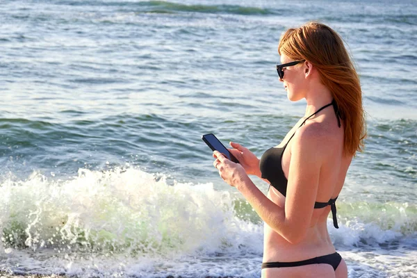 Милая улыбающаяся девушка пишет смс-ки со своего мобильного телефона на пляже. Приятный отдых Стоковое Изображение