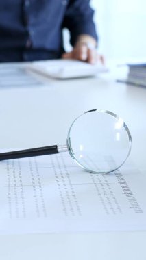 Magnifying glass resting on a financial report, with a businessman focused on a laptop in the blurred background, emphasizing financial analysis and due diligence. Audit and taxes theme in business