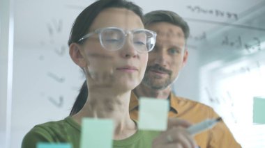 Businesswoman wearing glasses, writing on a glass board and organizing sticky notes, collaborates with adult businessman observing her strategy planning in a bright office.