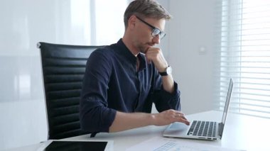 Businessman sitting at a sleek desk, working on a laptop and thinking in a bright, modern office filled with natural light. Business people concept.