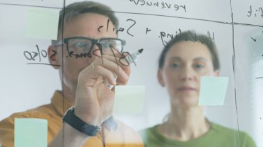 Two business professionals collaborating at a glass board, utilizing sticky notes to develop strategic plans while writing calculations and exchanging innovative ideas in a focused meeting