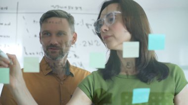 Two business colleagues brainstorming and planning together, using sticky notes and a marker on a glass board in a modern office, fostering teamwork and innovative ideas