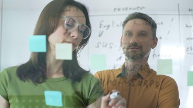 Businesswoman wearing glasses, writing on a glass board and organizing sticky notes, collaborates with a businessman observing her strategy planning in a bright office setting.