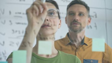 Businesswoman wearing glasses, writing on a glass board and organizing sticky notes, collaborates with adult businessman observing her strategy planning in a bright office.