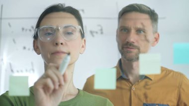Businesswoman wearing glasses, writing on a glass board and organizing sticky notes, collaborates with a businessman observing her strategy planning in a bright office setting