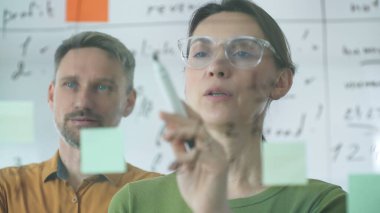 Businesswoman holding a marker while explaining strategy to a colleague, using a glass board filled with sticky notes during an engaging work meeting in a corporate office setting