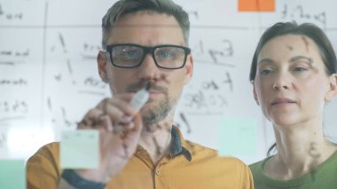 Focused business colleagues are discussing and planning strategy writing on a transparent whiteboard with colorful sticky notes during a collaborative working session