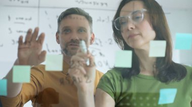 Businesswoman wearing glasses, writing on a glass board and organizing sticky notes, collaborates with a businessman observing her strategy planning in a bright office setting.