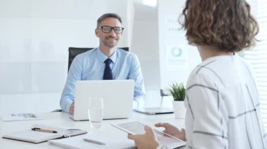 Two business colleagues collaborating on a project at a modern office desk, utilizing a laptop and tablet with documents and stationery nearby, creating a focused work environment