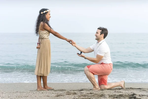 European man proposing to beautiful young woman with darker skin tone at the beach