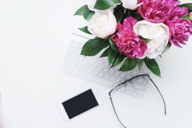 Klavye, not defteri, bardaklar, cep telefonu ve beyaz masa arkasında pembe ve beyaz şakayık çiçekleri olan işyeri. Düz yatış, üst görünüm